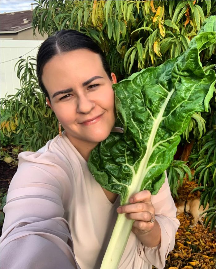 Paula Hardy standing in her home garden, smiling, holding a large Swiss chard leaf.