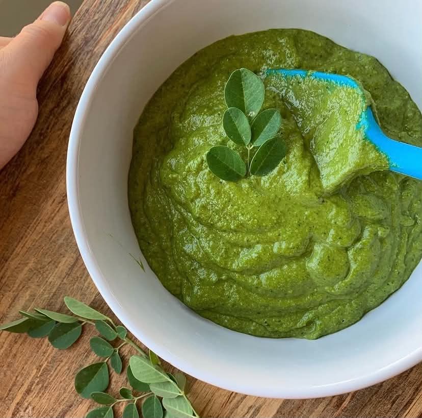 A hand-blended bowl of vibrant green moringa pesto, garnished with fresh moringa leaves, on a wood table.