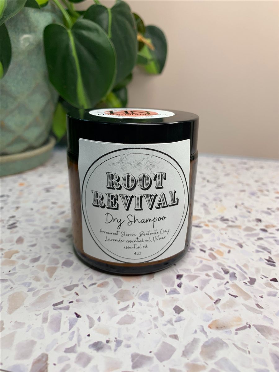 A jar of Root Revival dry shampoo, labeled, in front of a potted plant.