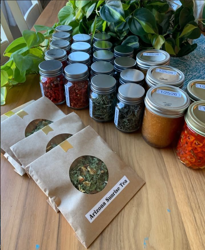 Kraft-paper Arizona Sunrise Tea packets and jars of dried herbs on a wood table with a leafy houseplant behind them.